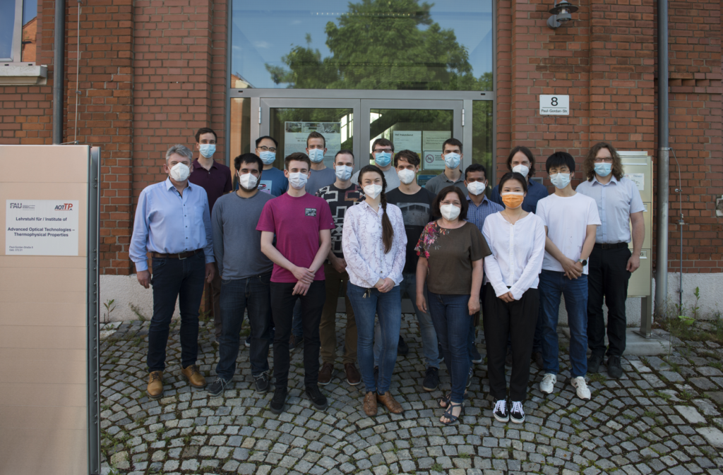 Photograph of AOT-TP team in June 2021; the group members wear masks and are standing in front of the entrance door of the institute