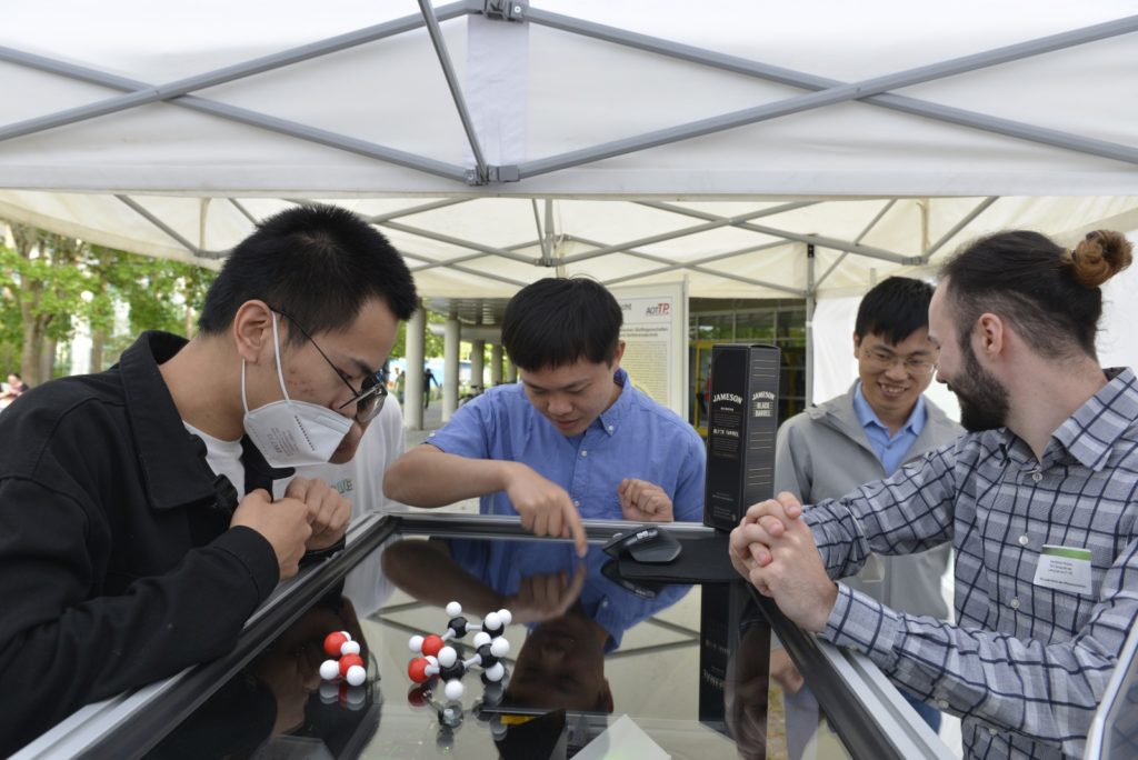 Photo of Taotao Zhan, Junwei Cui, and Maximilian Piszko showing the DLS setup to some interested guests
