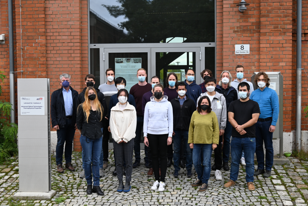 Photograph of AOT-TP team in September 2022; the group members wear masks and are standing in front of the entrance door of the institute